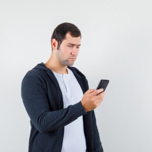 A young man sitting on a couch holding his phone with a worried expression, avoiding answering a call due to social anxiety.