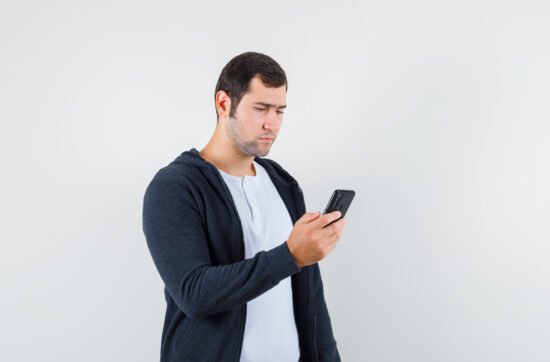 A young man sitting on a couch holding his phone with a worried expression, avoiding answering a call due to social anxiety.