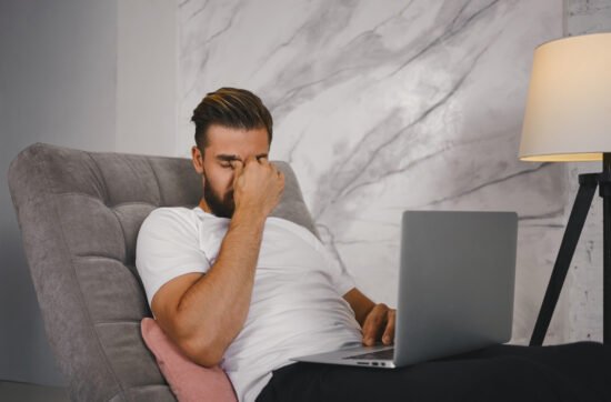 A man sitting on his couch late on Sunday evening, looking stressed while staring at his laptop, dreading Monday morning.