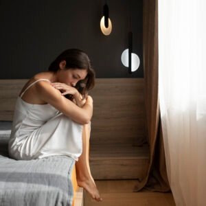 A young woman sitting on the edge of her bed in the morning, looking down with exhaustion and sadness, struggling to start the day.