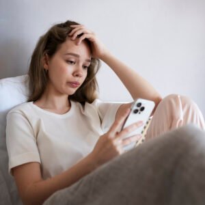 A woman sitting alone on a couch at home, looking at her phone with sadness after cancelling plans.