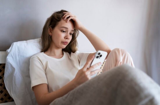 A woman sitting alone on a couch at home, looking at her phone with sadness after cancelling plans.