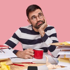 A young man sitting at a cluttered desk with his head in his hands, surrounded by unfinished tasks and papers.
