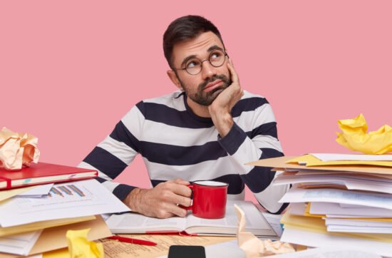 A young man sitting at a cluttered desk with his head in his hands, surrounded by unfinished tasks and papers.