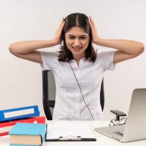 A young woman sitting in an office wearing noise-cancelling headphones, closing her eyes with relief as she blocks out distracting sounds.