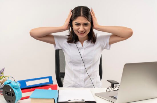 A young woman sitting in an office wearing noise-cancelling headphones, closing her eyes with relief as she blocks out distracting sounds.