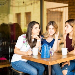 A young woman sitting quietly at a table with a group of friends laughing and talking around her, looking thoughtful and slightly withdrawn.