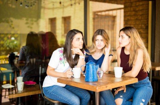 A young woman sitting quietly at a table with a group of friends laughing and talking around her, looking thoughtful and slightly withdrawn.
