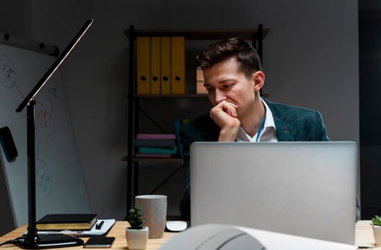 A young professional man sitting at his desk with a laptop open, looking anxious despite appearing successful at work.