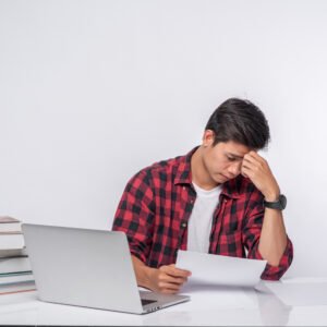 A young man sitting at a desk surrounded by papers and a laptop, looking frustrated as he revises his work again and again.