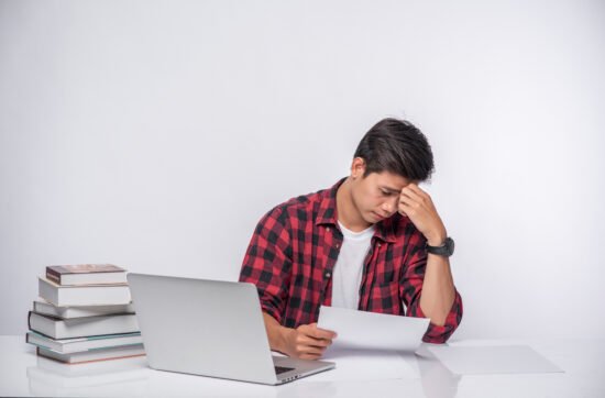 A young man sitting at a desk surrounded by papers and a laptop, looking frustrated as he revises his work again and again.