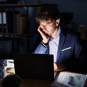 A man sitting at his desk late at night with papers and a laptop in front of him, looking exhausted as his to-do list grows longer.