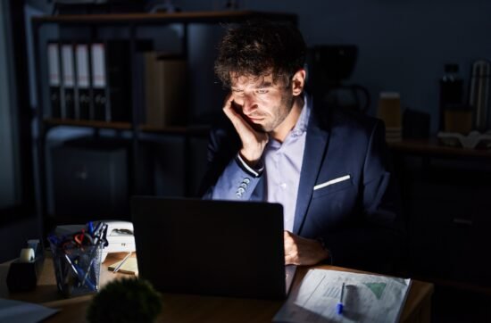 A man sitting at his desk late at night with papers and a laptop in front of him, looking exhausted as his to-do list grows longer.