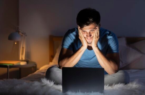 A man sitting on his couch on Sunday evening, staring at his laptop with a worried expression as he thinks about the work week ahead.