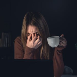 A woman sitting at her dining table late at night with her head resting on her arms, surrounded by papers and her phone buzzing with new messages.