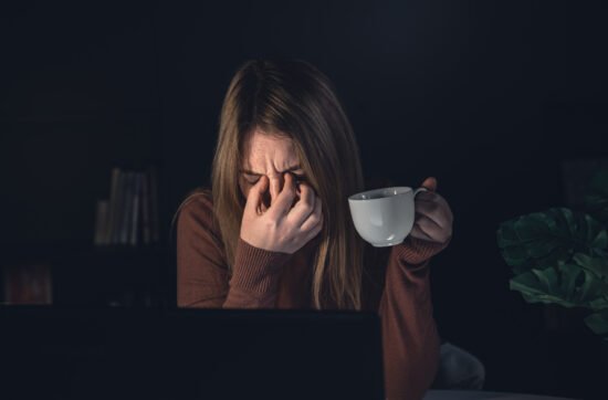 A woman sitting at her dining table late at night with her head resting on her arms, surrounded by papers and her phone buzzing with new messages.