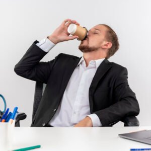 A man sitting at his desk with an empty coffee cup in his hand, leaning back with a tired expression despite drinking multiple cups.