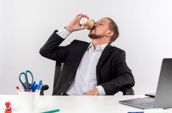 A man sitting at his desk with an empty coffee cup in his hand, leaning back with a tired expression despite drinking multiple cups.